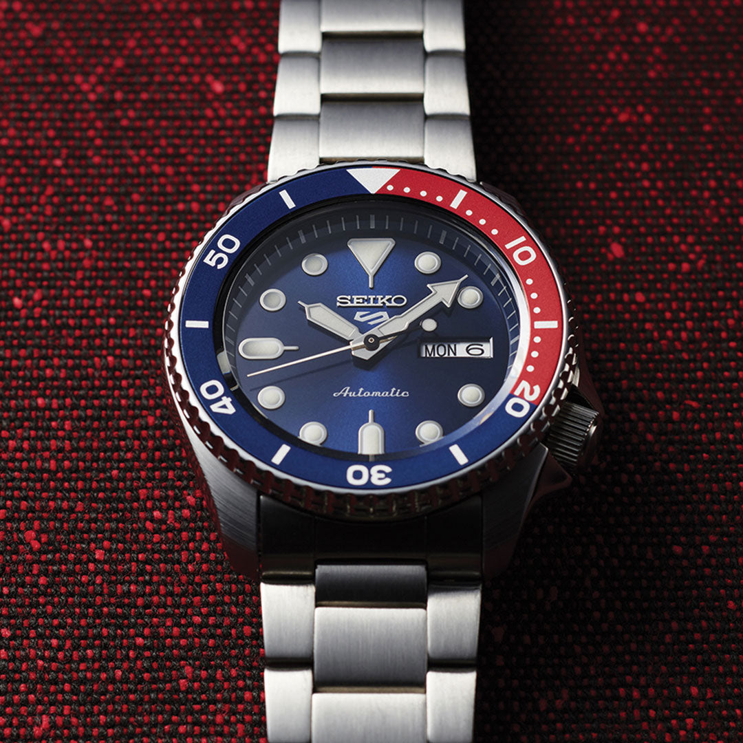 The Seiko 5 Sports SKX Series Automatic 42.5mm Watch by Seiko boasts a stainless steel band and case with an eye-catching blue and red bezel on a textured red background. The dial showcases the day and date, featuring bold, luminescent hour markers and hands.