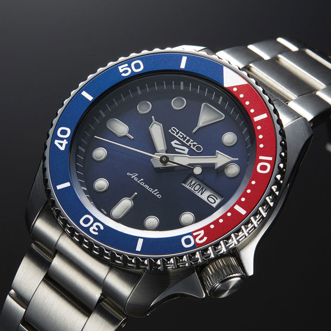 A detailed view of the Seiko 5 Sports SKX Series Automatic 42.5mm Watch highlights its blue and red bezel and sturdy stainless steel case. The watch features a blue dial with luminescent hour markers, silver hands, and a day-date display reading "MON 6," all driven by automatic movement. The bezel is decorated with minute markings for added precision.