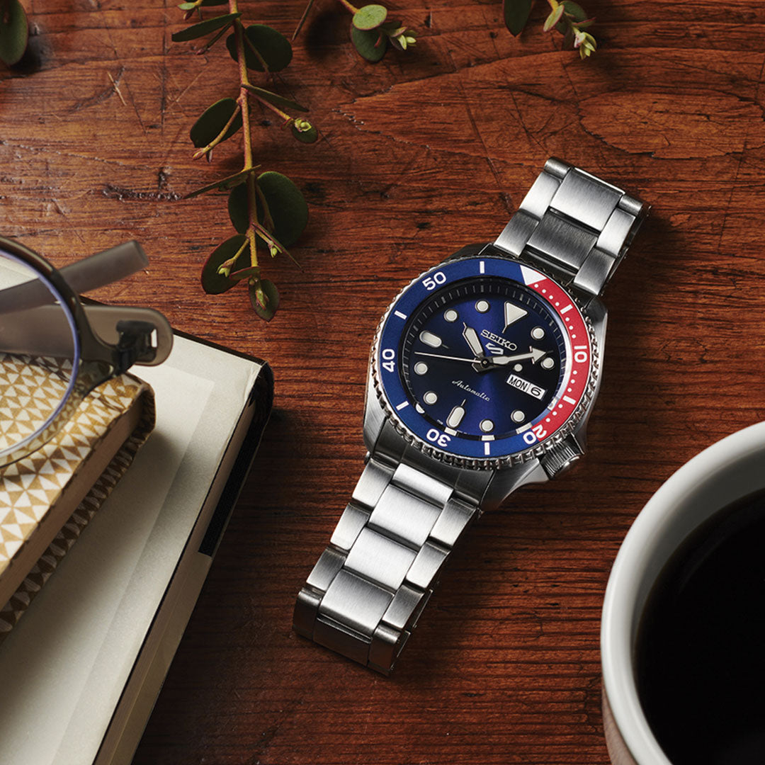 A Seiko 5 Sports SKX Series Automatic 42.5mm Watch, featuring a stainless steel case and automatic movement, is displayed on a wooden table beside a pair of glasses, a notebook, and a cup of coffee. The watch's blue and red bezel offers an elegant contrast to its dark blue dial, with small green plant leaves scattered nearby.