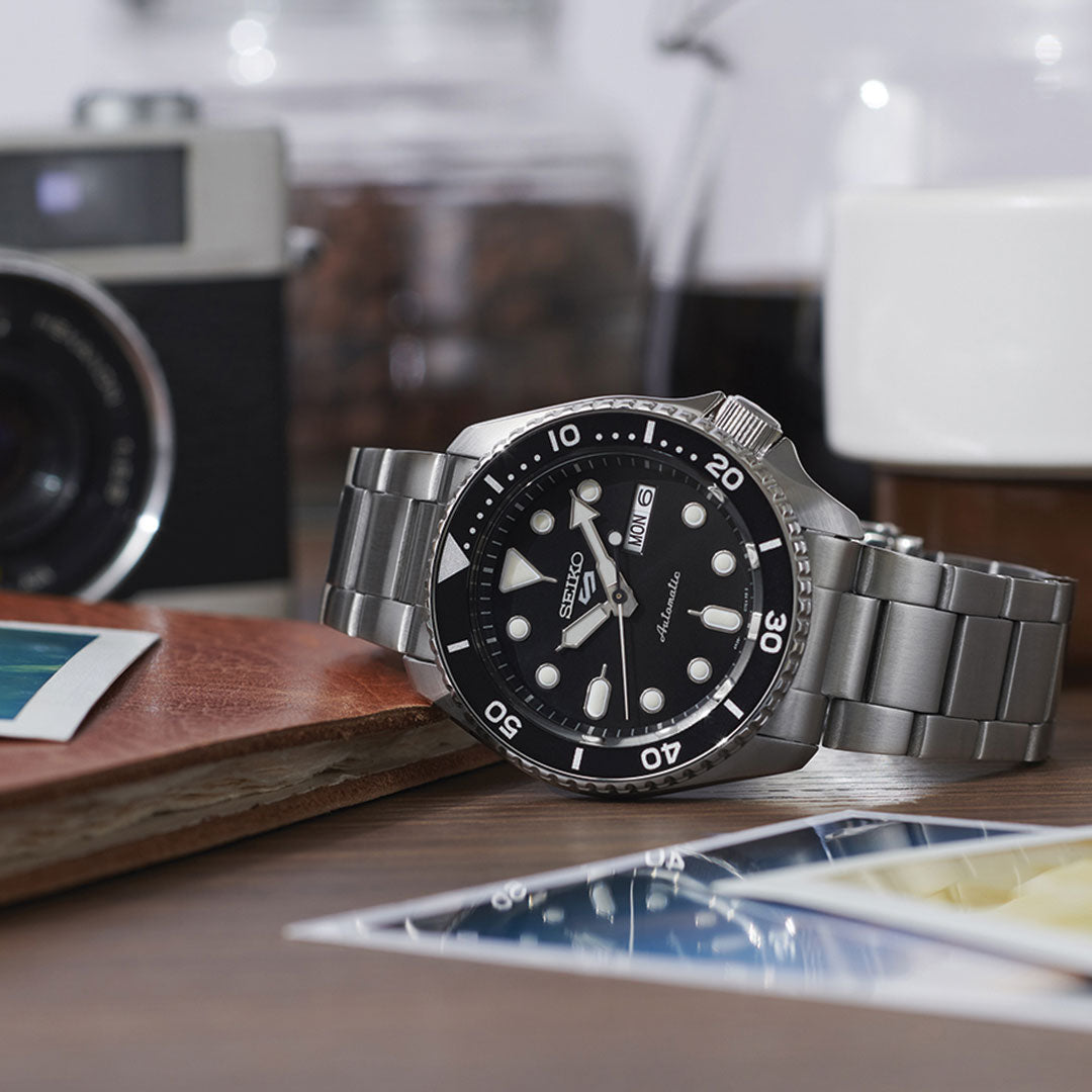 A Seiko 5 Sports SKX Series Automatic 42.5mm watch with a black dial and bezel rests on wood near a leather notebook, instant photos, and a vintage camera in the background.