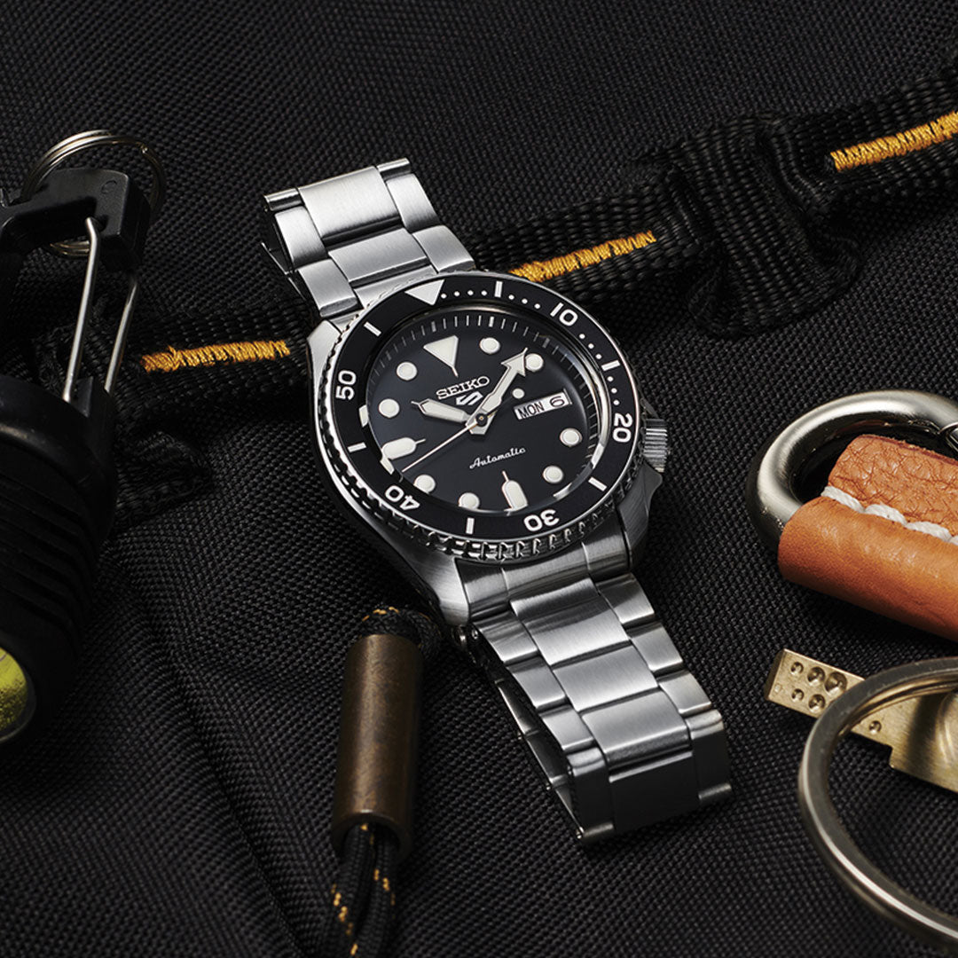 The Seiko 5 Sports SKX Series Automatic 42.5mm Watch by Seiko, featuring a stainless steel case with black dial and bezel, is displayed on dark fabric alongside outdoor gear like a flashlight, rope, and leather key holder.