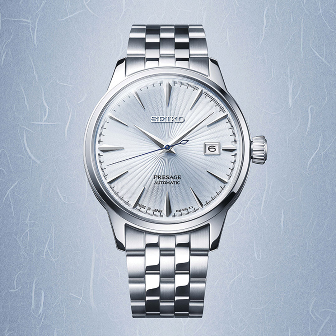 The Seiko Presage Cocktail Time Automatic 40.5mm features a silver case, textured light blue dial, date at 3 o’clock, and metal link bracelet, showcased against a subtle blue patterned backdrop.