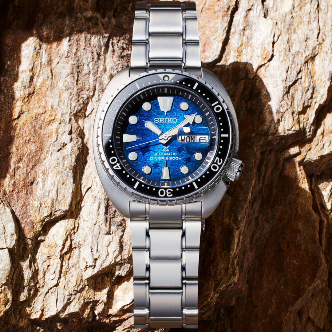 The Seiko Prospex Sea Save The Ocean Special Edition Automatic With Manual Winding 45mm Watch, featuring a silver case and a blue patterned dial, is displayed against a textured rock background. The watch face shows the time 10:08 and includes a day-date window at the three o'clock position. Equipped with an automatic Caliber 4R36, its black bezel is detailed with white markings to ensure precision timing.