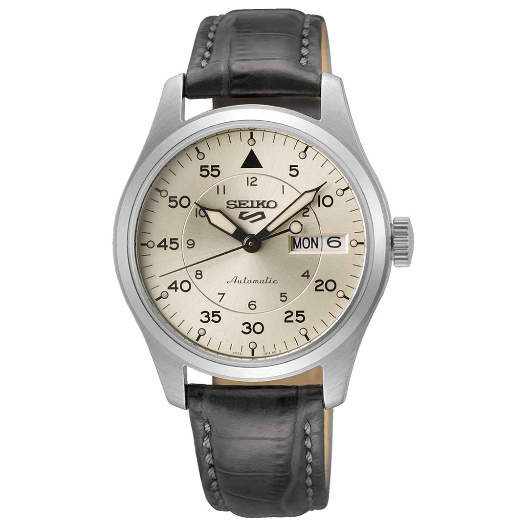 A Seiko 5 Sports Field Series Automatic 36.37mm Watch features a stainless steel silver case and a black leather strap. It has a beige dial with Arabic numerals at every five-minute interval, a day/date display window at 3 o'clock, and black hour and minute hands on this automatic timepiece by Seiko.