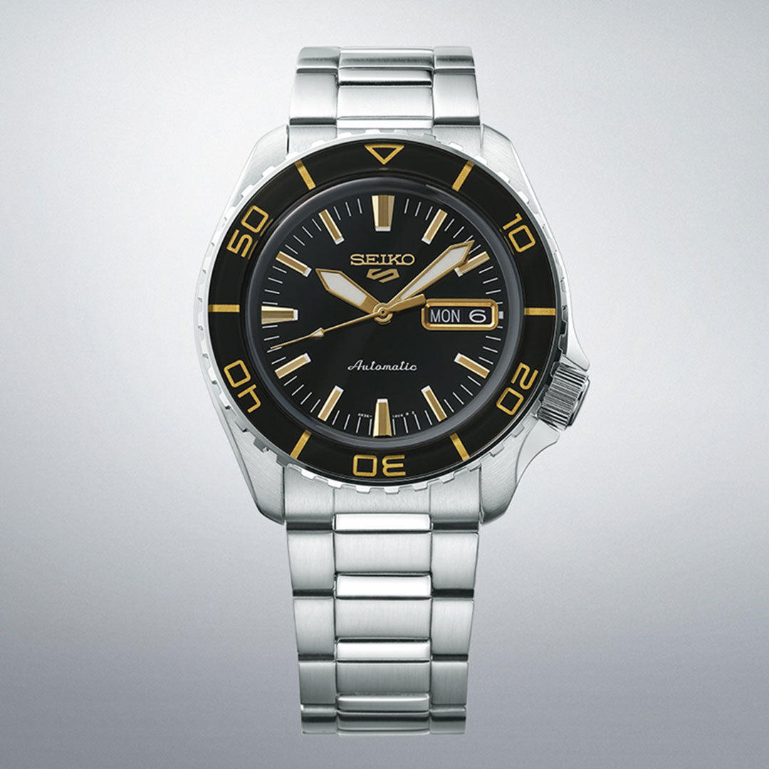 Introducing the Seiko 5 Sports SKX Series Automatic 42.5mm Watch. This stainless steel timepiece boasts a silver design complemented by a black face and sophisticated gold accents. Featuring a day-date display on the right side, it also includes a metal link bracelet for added style. The bezel is elegantly detailed with gold numbers at intervals, enhancing its refined appearance.