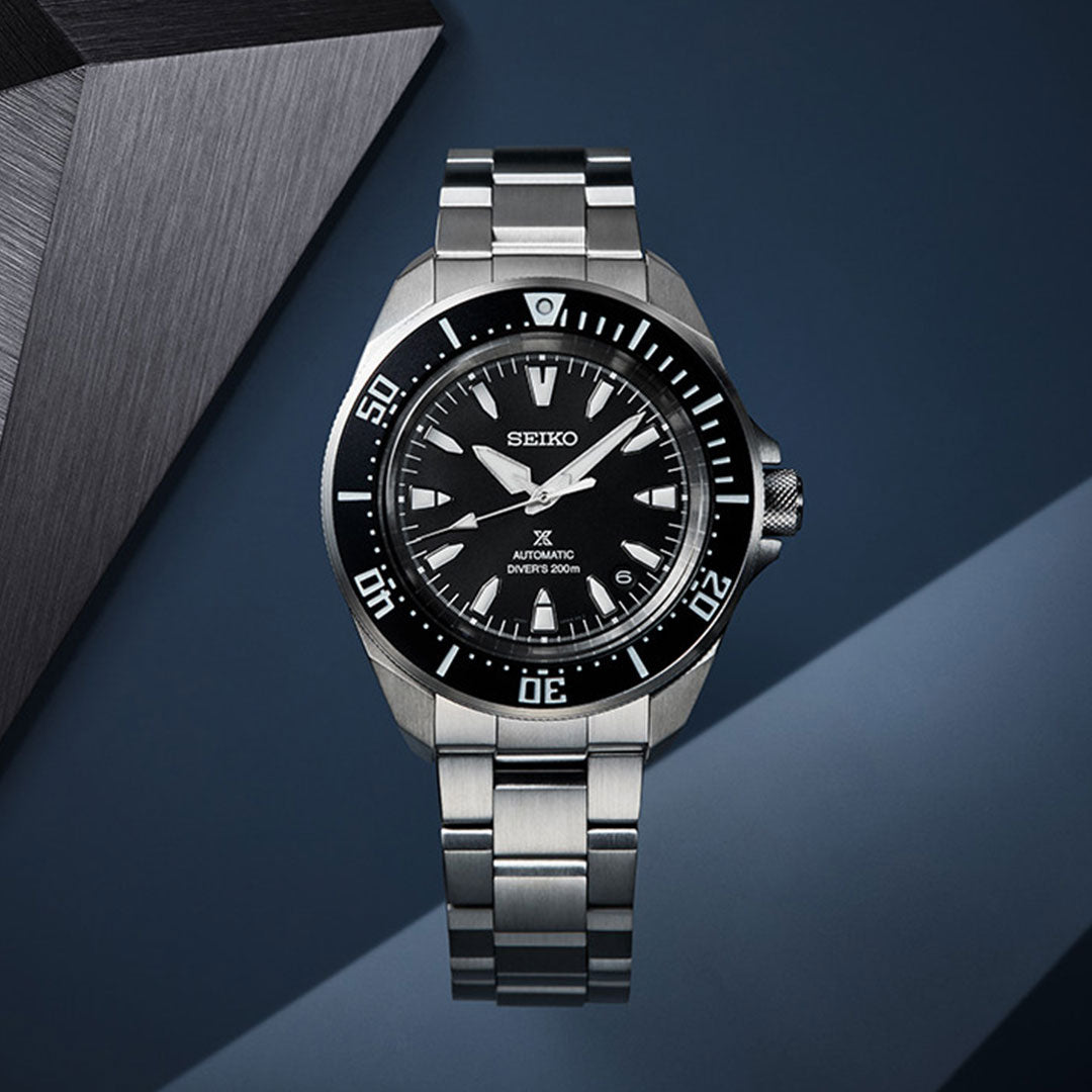 A Seiko Prospex Diver's Automatic 42mm Watch with a stainless steel bracelet, black dial, and rotating bezel is showcased against a contemporary geometric backdrop in shades of grey and blue. The Seiko automatic movement ensures precision while its luminescent hands and markers enhance functionality.