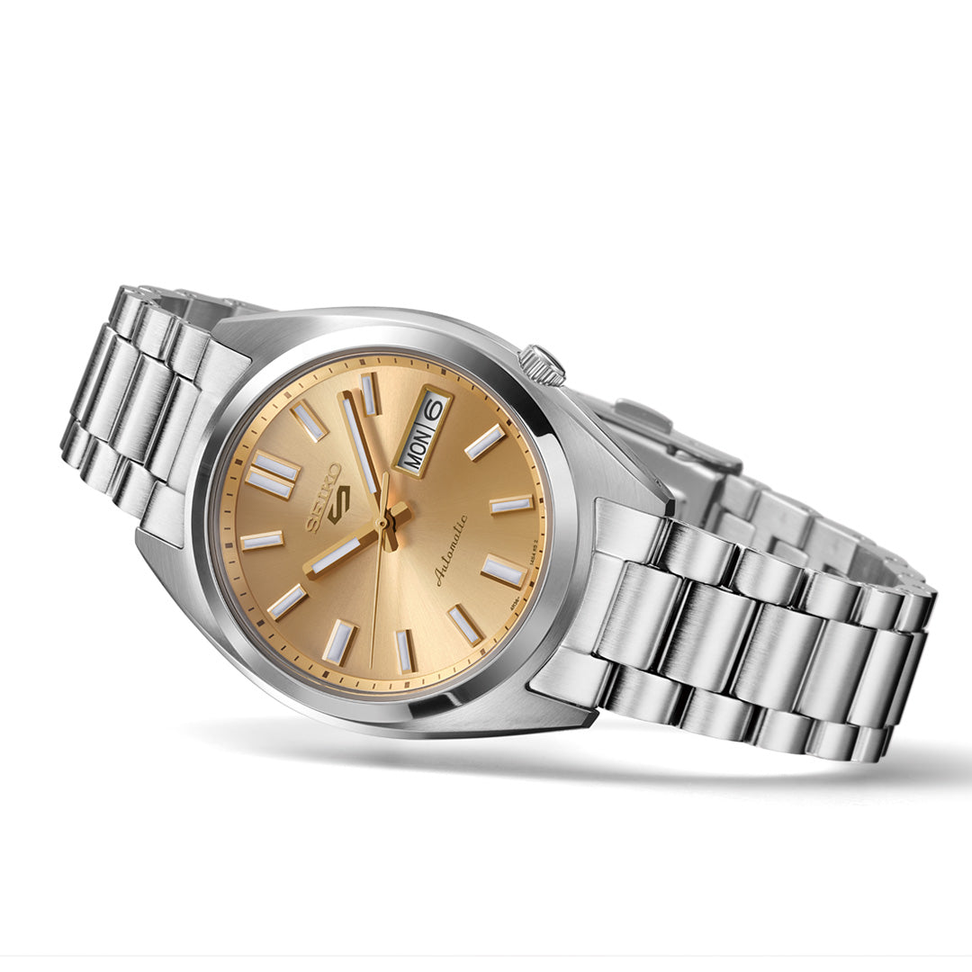 A Seiko 5 Sports SNXS Series Automatic 37.4mm watch by Seiko, featuring a gold dial with metallic hour markers and a day-date display, is displayed on a stainless steel bracelet against a white background.