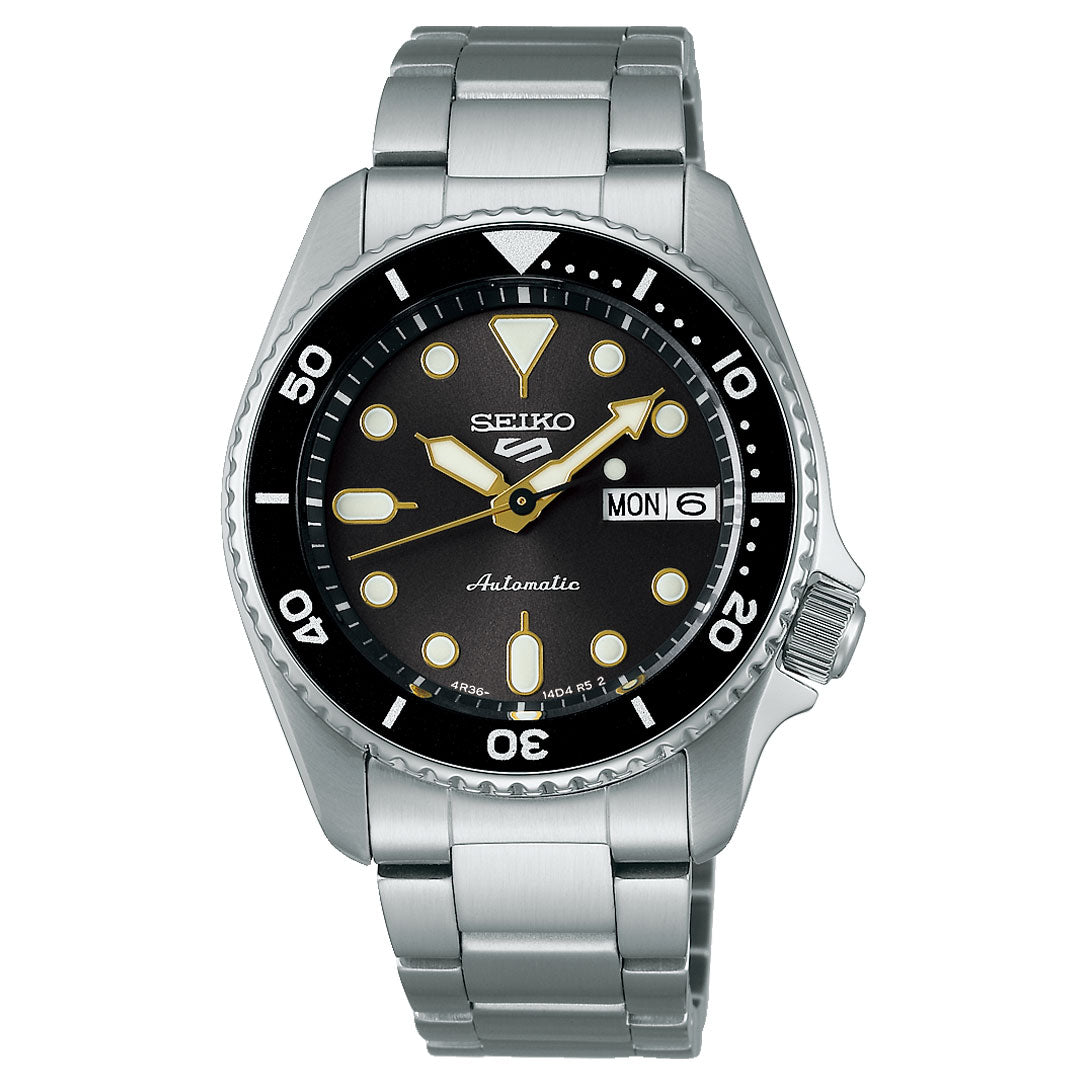 The Seiko 5 Sports SKX Series Automatic 38mm watch features a stainless steel case, black bezel and dial, white and gold markers, day-date display set to Monday the 6th, and a silver metal link band.
