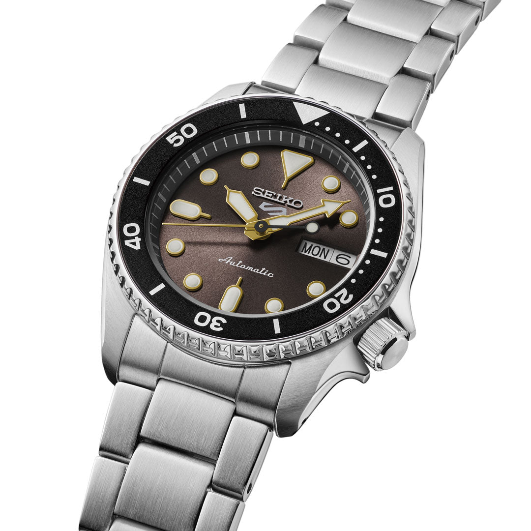 The Seiko 5 Sports SKX Series Automatic 38mm watch features a stainless steel case, brown gradient dial with yellow markers, day-date at 3 o’clock, black bezel with white markings, and a silver metal bracelet.