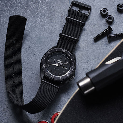 A Seiko 5 Sports SKX Series Automatic 42.5mm watch with a fabric strap rests on a grey surface near skateboard parts, bolts, and nuts, with a skateboard partially visible in the lower right corner.