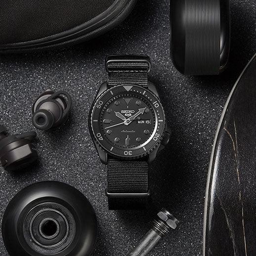 The Seiko 5 Sports SKX Series Automatic with Manual Winding 42.5mm watch by Seiko, featuring a black case and fabric strap, rests on a dark textured surface beside black skateboard wheels, earbuds, and a protective case.