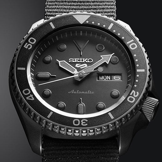Close-up of the Seiko 5 Sports SKX Series Automatic 42.5mm watch, featuring a dark dial, bold white luminous hands and markers, day-date display, textured bezel, and fabric strap.