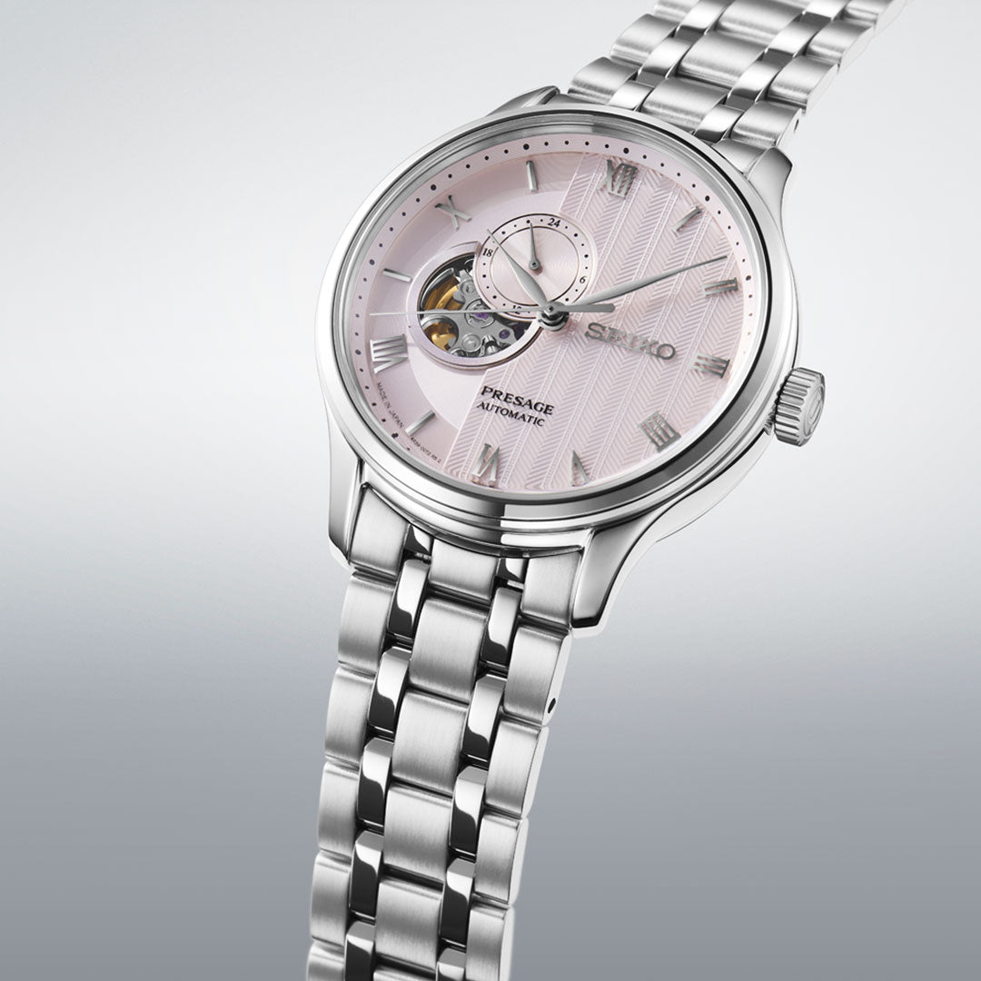 A Seiko Presage Japanese Garden Automatic 41.8mm watch with a stainless steel band and light pink textured dial, open-heart display, and subdial is photographed against a light gray background.