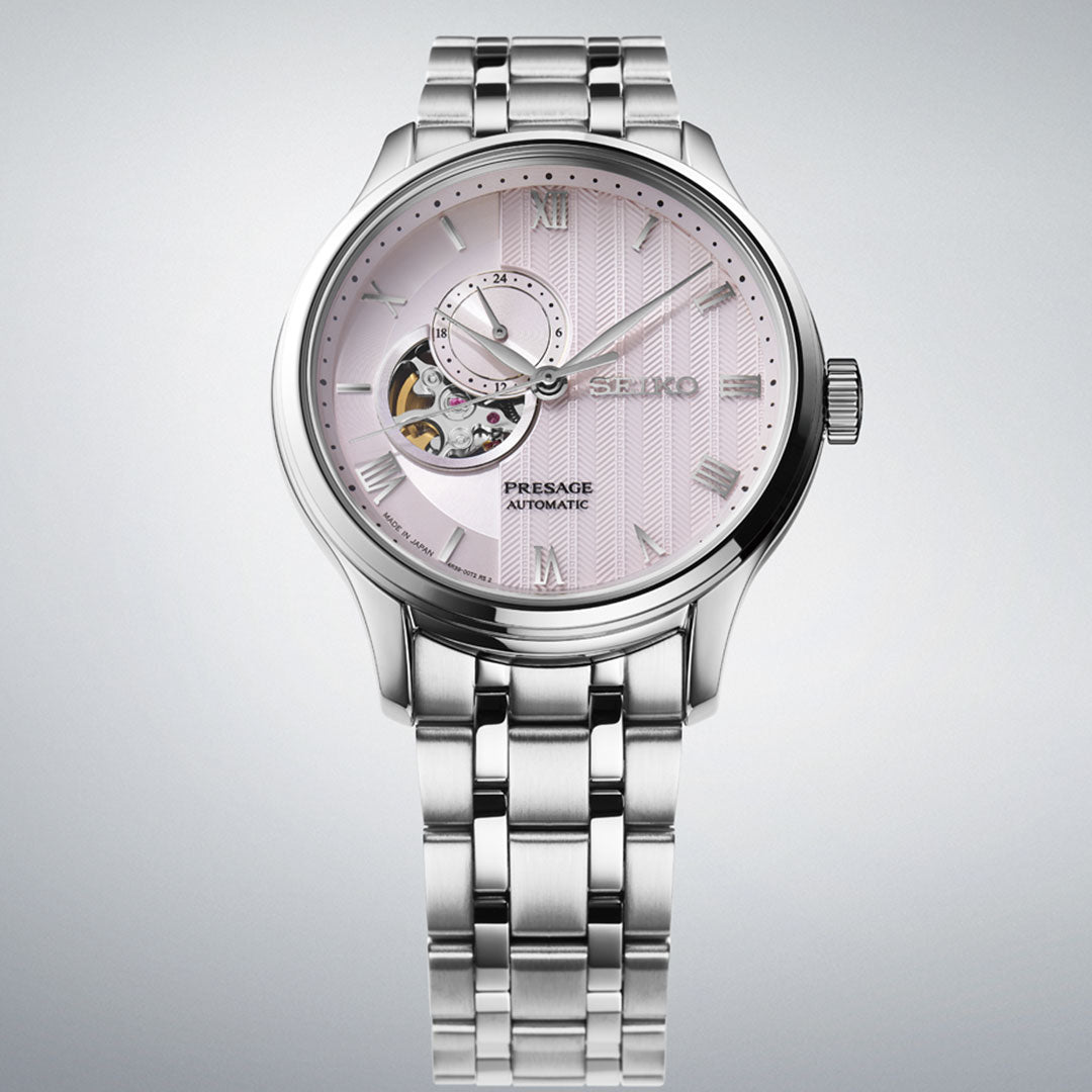 The Seiko Presage Japanese Garden Automatic, a 41.8mm watch, features a light pink dial with Roman numerals, an open-heart design showing the movement, a small sub-dial, and a silver metal band.