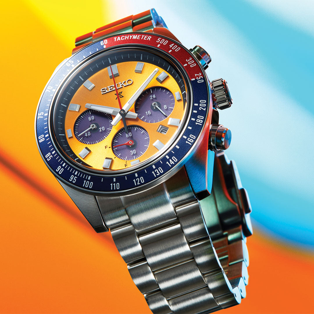 The Seiko Prospex Speedtimer Solar 41.4mm Watch is a vibrant wristwatch featuring a yellow face and blue tachymeter bezel. This solar-powered timepiece boasts three sub-dials, silver hands, and a durable design, all complemented by a stainless steel bracelet. The background beautifully blends shades of orange and blue.