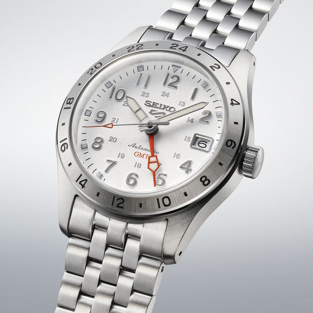 The Seiko 5 Sports Field Series Automatic 39.4mm Watch features a white dial with silver markers, black numerals, a red GMT hand, and a date window at 3 o’clock on a stainless steel bracelet, shown against a light background.
