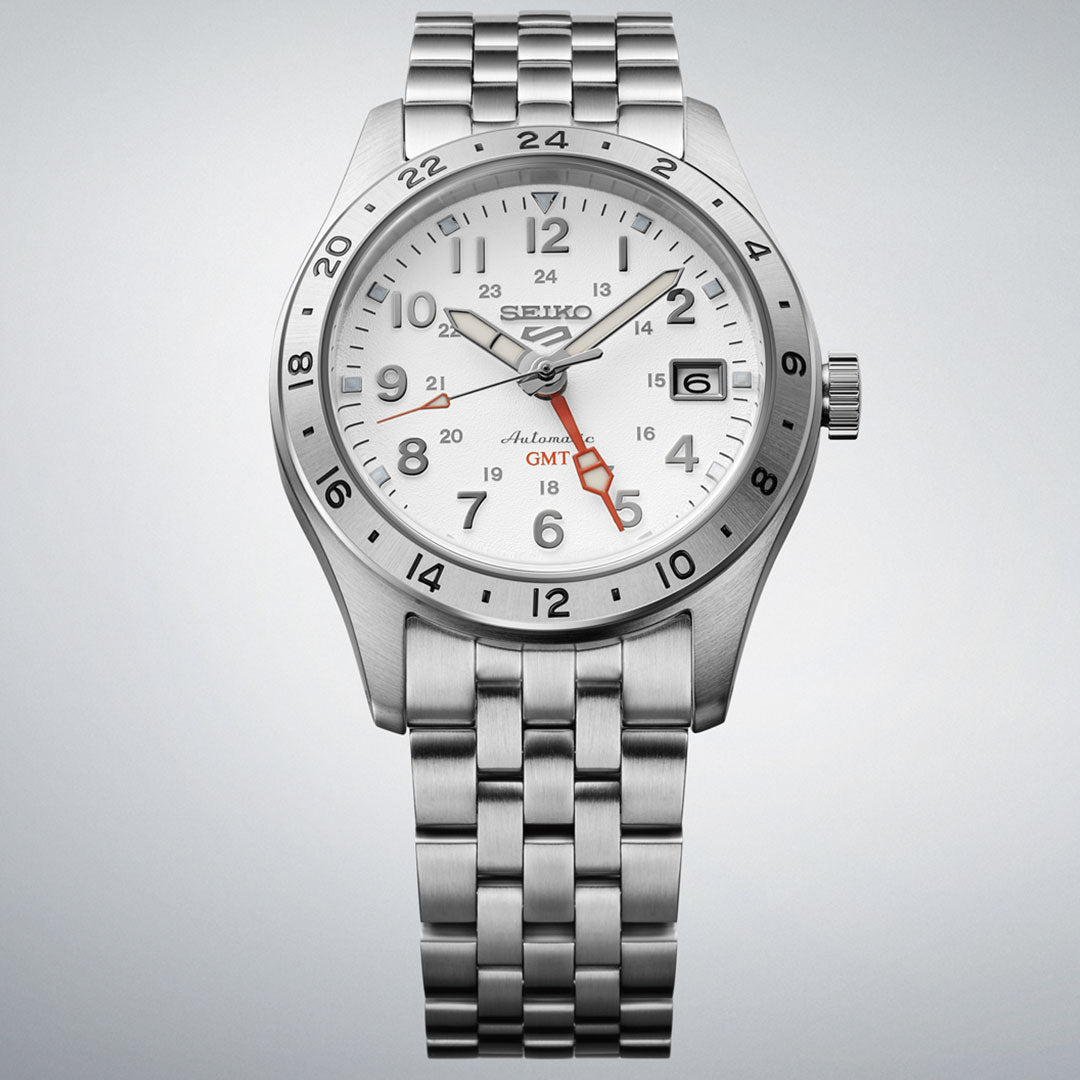 The Seiko 5 Sports Field Series Automatic 39.4mm Watch features a stainless steel case, white dial, black markers, red GMT hand, date at 3 o’clock, and metal link bracelet, shown on a light gray background.