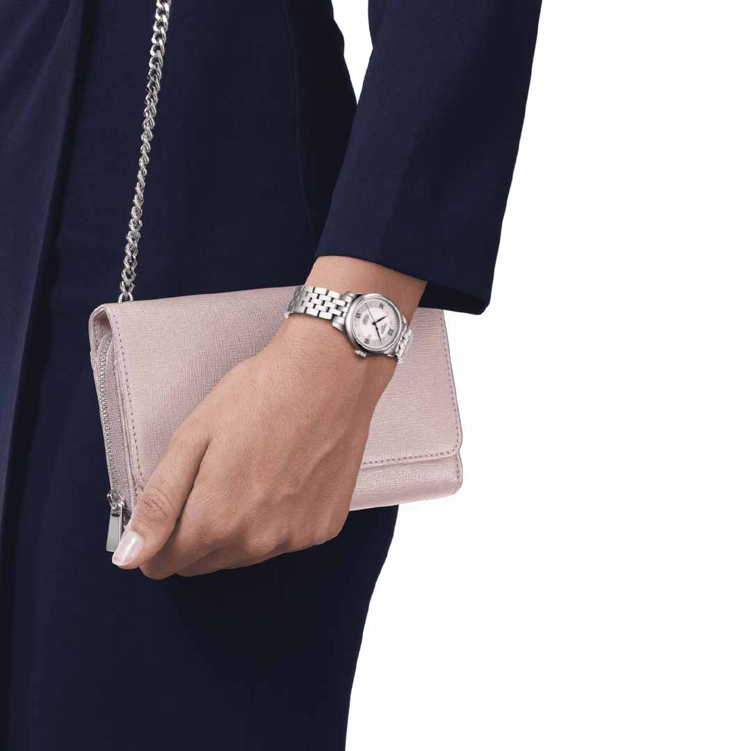 Dressed in a dark blue ensemble, a person holds a light pink clutch purse adorned with a silver chain. On their left wrist is the elegant Tissot Le Locle Automatic 29mm Watch 20th Anniversary Edition, showcasing its classic round face that effortlessly complements their sleek style.