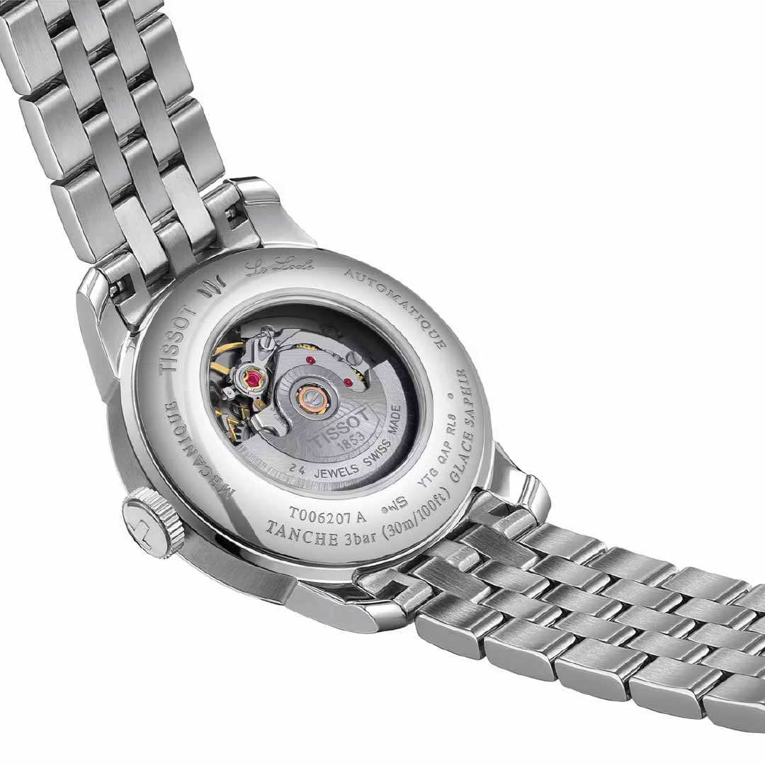 The image showcases the back view of a Tissot Le Locle Automatic 29mm Watch 20th Anniversary edition, featuring a stainless steel band. The transparent case back elegantly reveals the intricate mechanical movement inside, highlighting visible gears and displaying the inscription "Tissot 1853" around the edge.