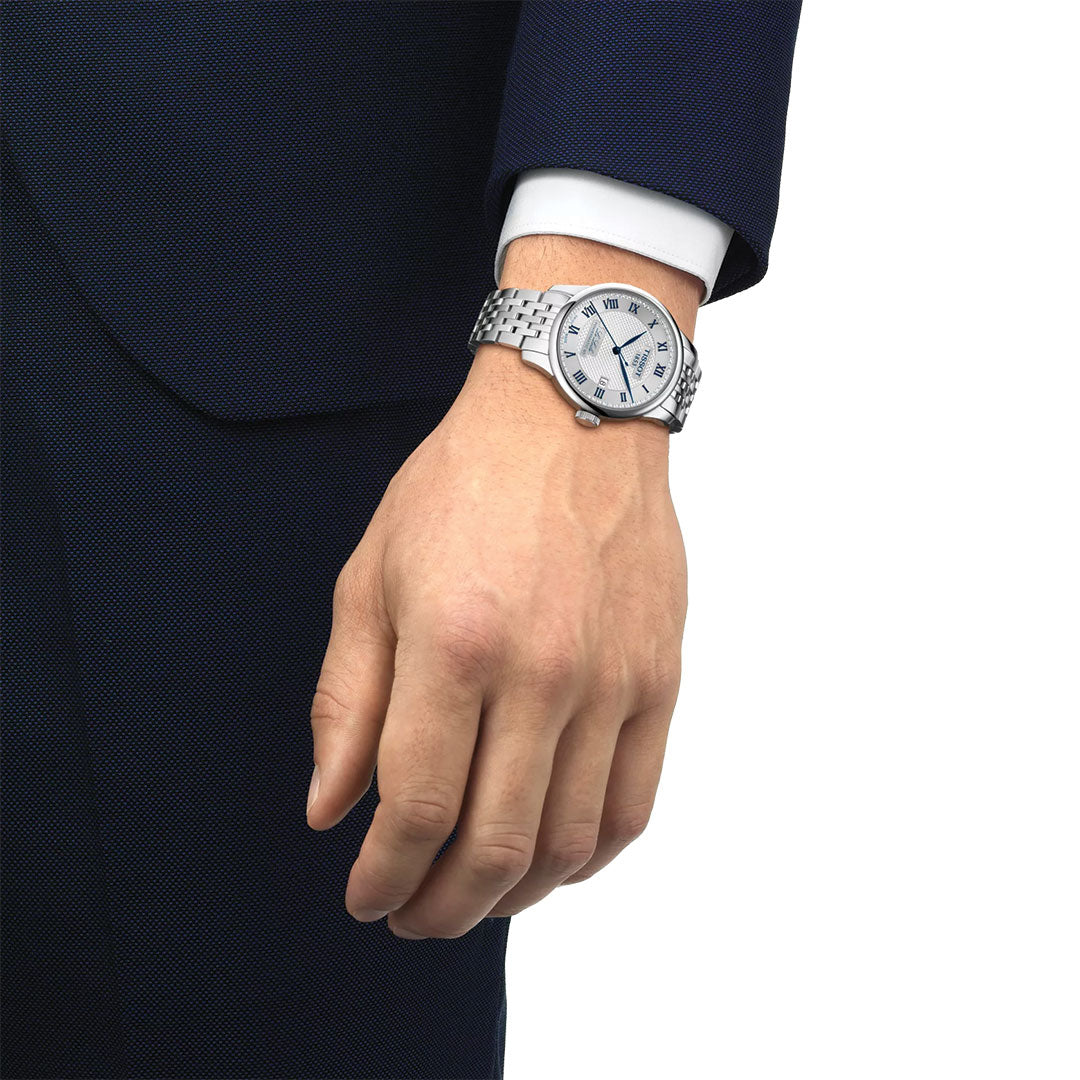 A person in a navy blue suit and white shirt wears the Tissot Le Locle 20th Anniversary Powermatic 80 Automatic 39.3mm Watch, featuring a silver metal band, white face, and Roman numerals on their relaxed left wrist.