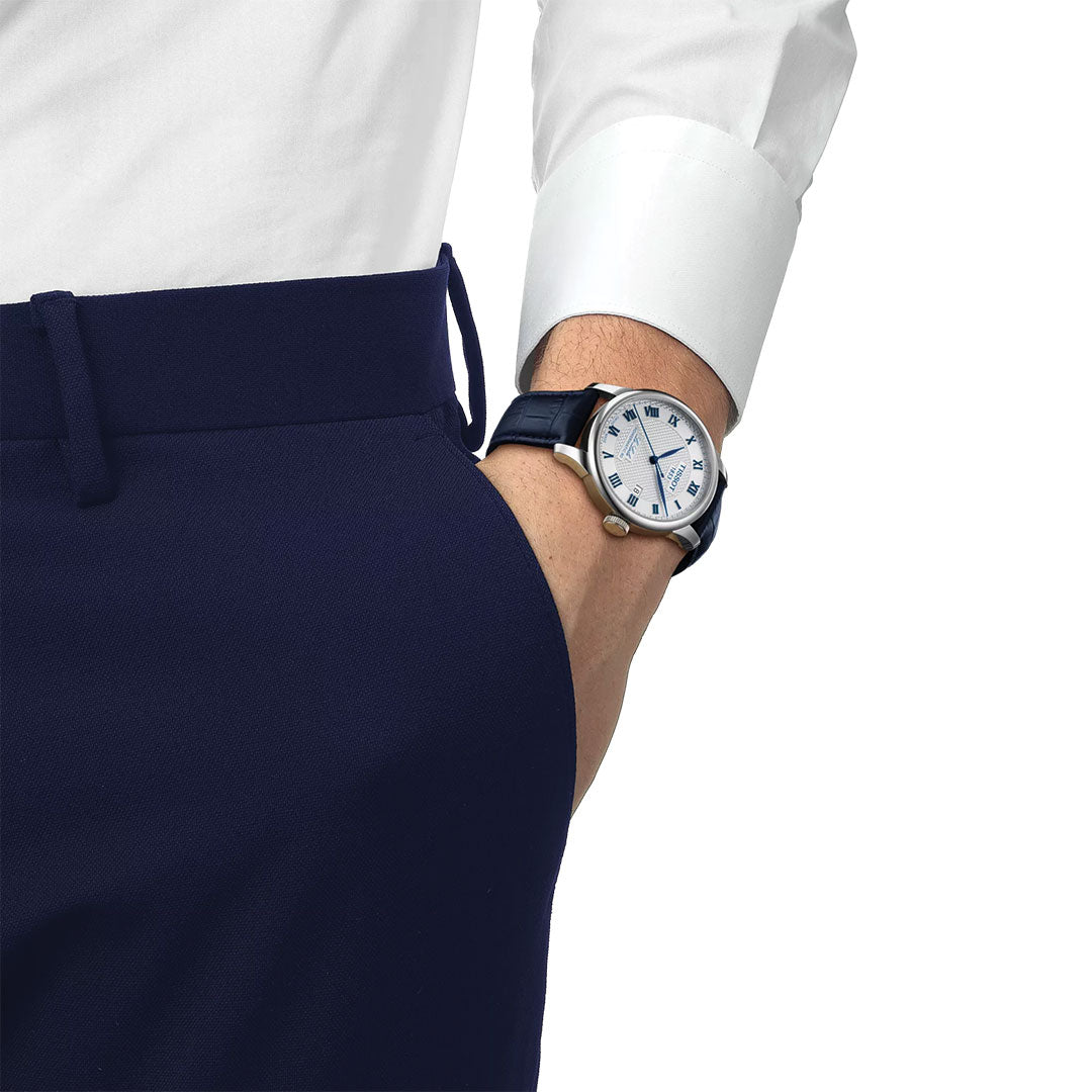 A person in a white dress shirt and navy pants showcases the Tissot Le Locle 20th Anniversary Powermatic 80 Automatic 39.3mm Watch by Tissot, featuring an 80-hour power reserve, blue strap, and white dial with blue Roman numerals.