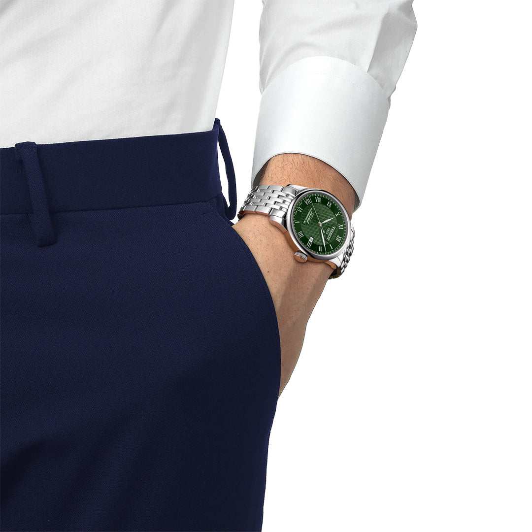 A person in navy trousers and a white shirt has their hand in their pocket, showing a Tissot Le Locle Powermatic 80 Automatic 39.3mm watch with a green face on their left wrist, reflecting Tissot’s Swiss heritage.