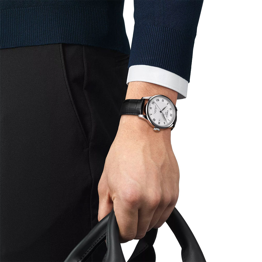 A person in a dark blue sweater and black pants holds a black leather bag. On their left wrist is a Tissot Le Locle Powermatic 80 Automatic 39.3mm watch with a white face and scratch-resistant sapphire crystal.