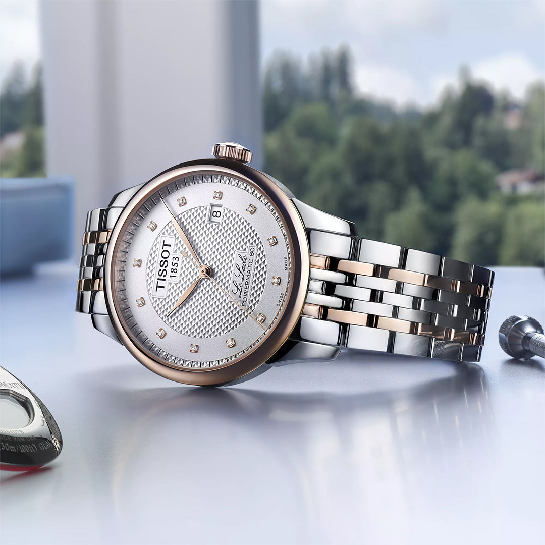 The Tissot Le Locle Powermatic 80 Automatic 39.3mm Watch features a silver and rose gold metal band, a textured silver dial with date window, and is elegantly displayed on a reflective surface against blurred greenery and sky.
