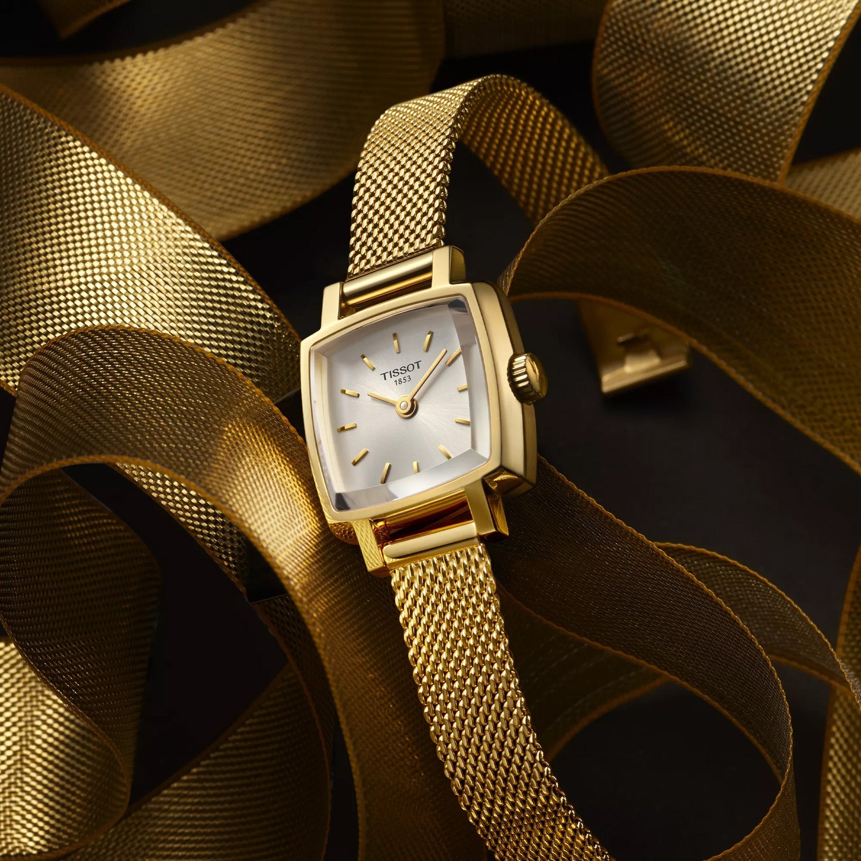 A Tissot Lovely Square Quartz 20 x 20mm watch with a gold mesh band is displayed on a black surface, exuding timeless glamour. Gold ribbons artistically intertwined around it highlight the elegance of ladies' watches.