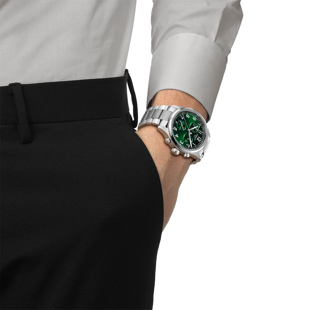 A person in a white dress shirt and black pants shows off a Tissot CHRONO L Quartz 42mm watch with a green face and white numerals, highlighting refined Swiss craftsmanship from Tissot.