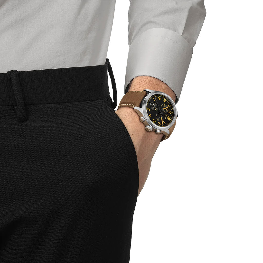 A person in black pants and a white dress shirt, hand in pocket, showcases the Tissot CHRONO L Quartz 42mm Watch by Tissot—with a brown leather strap and black dial featuring yellow accents, made with Swiss technology.