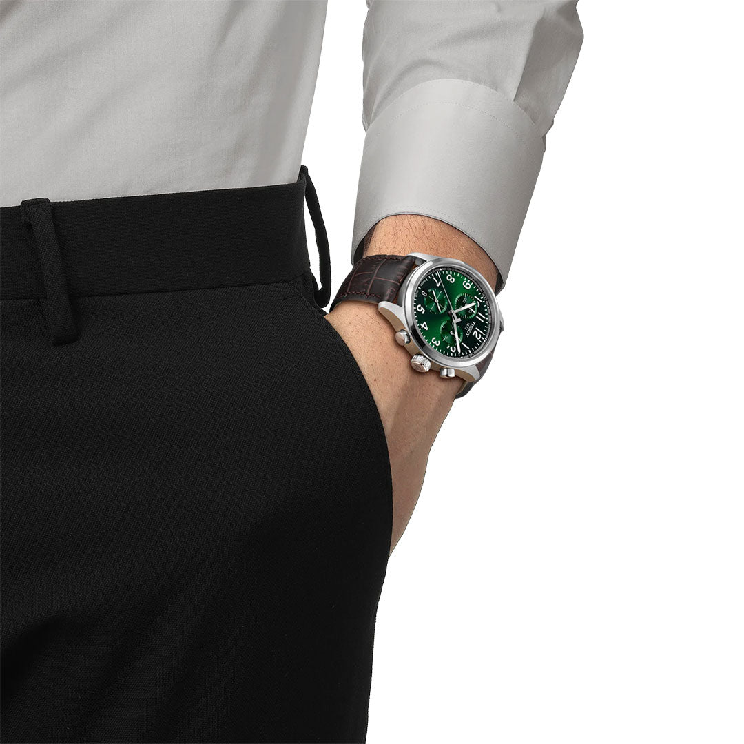 A person in a white dress shirt and black pants, hand in pocket, wears a Tissot CHRONO L Quartz 42mm watch with a green dial and brown leather strap on their wrist.