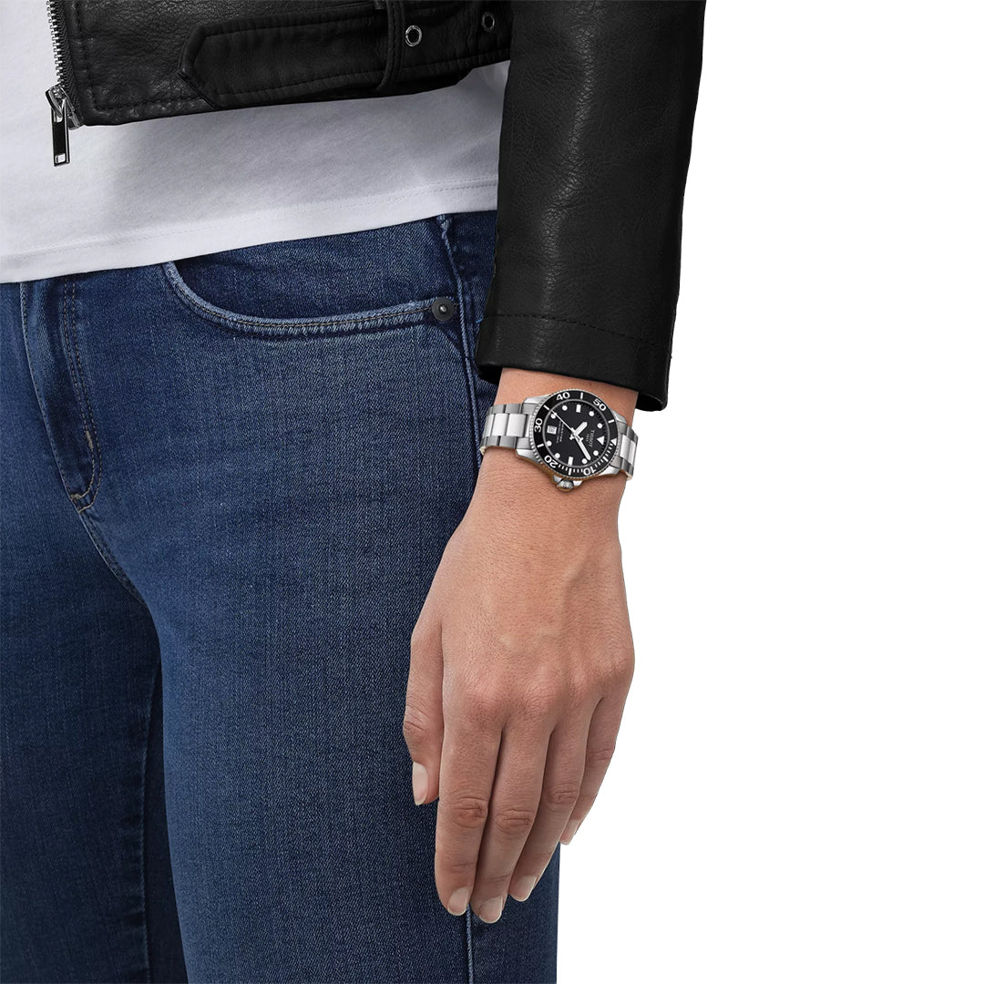 Wearing a black leather jacket, white shirt, and blue jeans, a person shows off a Tissot Seastar 1000 Quartz 36mm Watch by Tissot with a stainless steel case and black face on their left wrist. Only the lower torso and left hand are visible.