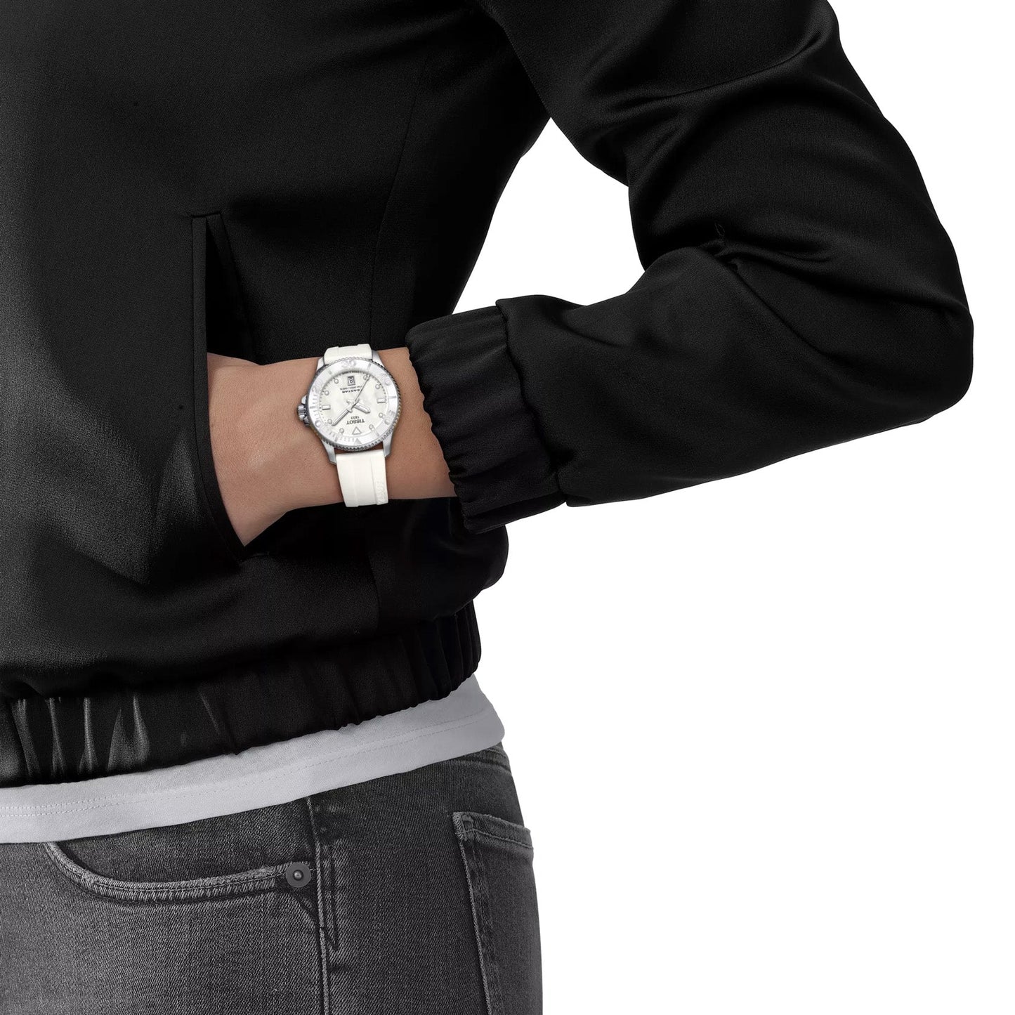 A person dressed in a black jacket poses with one hand in the pocket, showcasing a Tissot Seastar 1000 Quartz 36mm wristwatch. The watch features a white face that complements its 316L stainless steel body. The lower part of the jacket is visible alongside gray jeans and the hem of a white shirt.