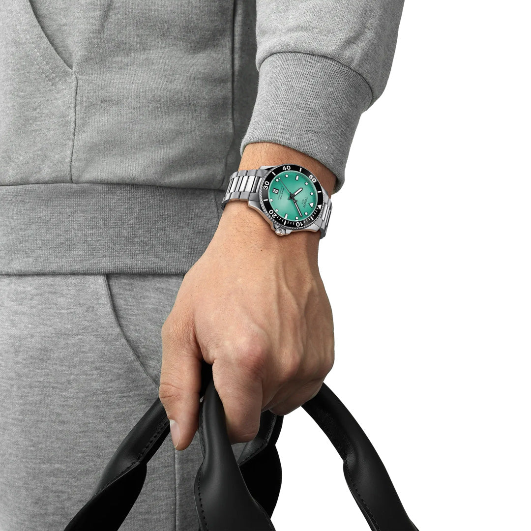 A person in a gray tracksuit, holding black handles, wears a Tissot Seastar 1000 Quartz 40mm Watch with a green face on their left wrist—a timepiece from Tissot known for Swiss craftsmanship and an impressive 300m water resistance.