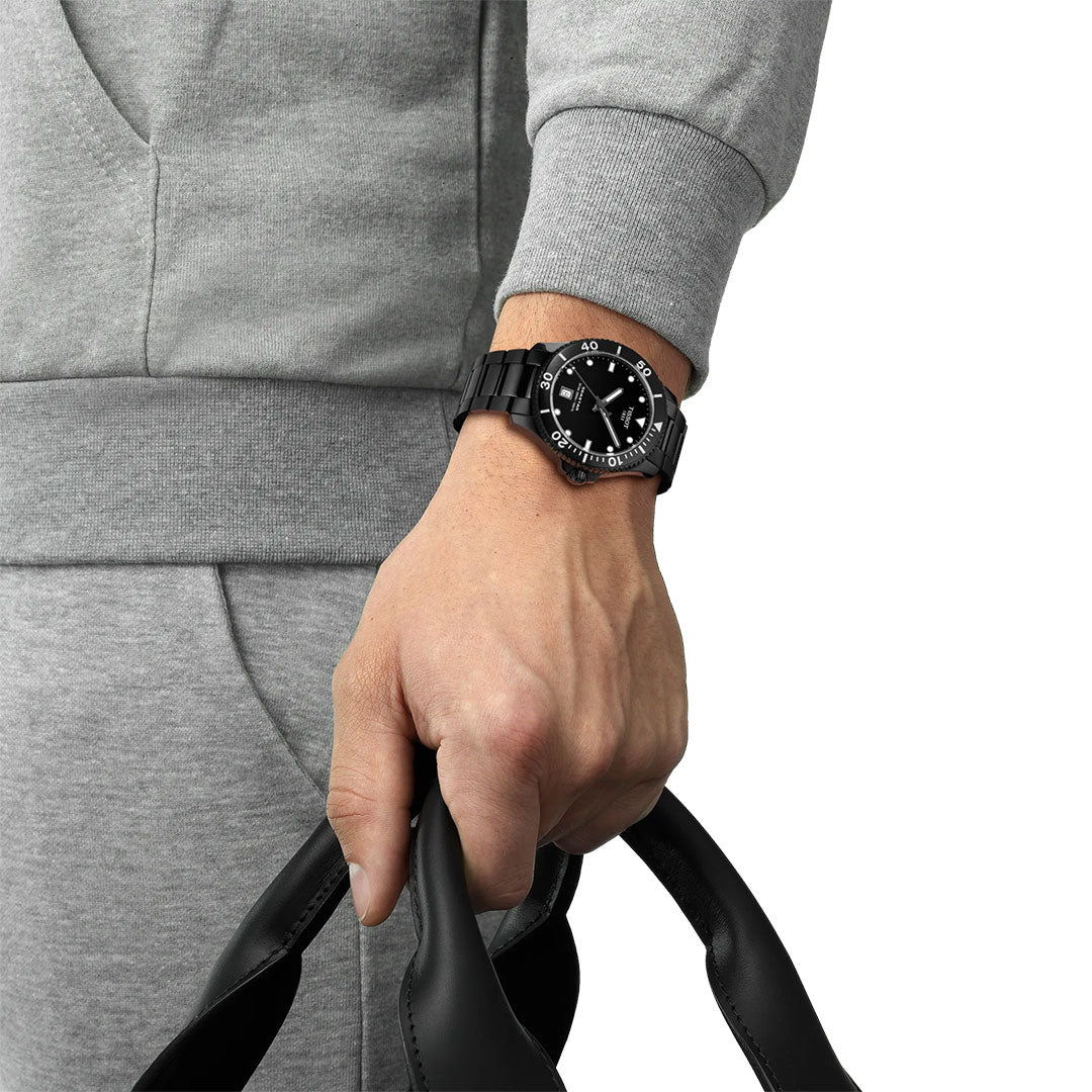 A person in a grey tracksuit holds black bag handles, wearing the Tissot Seastar 1000 Quartz 40mm Watch—a Swiss-made timepiece with a round black face, white markings, and water resistance from the brand Tissot.