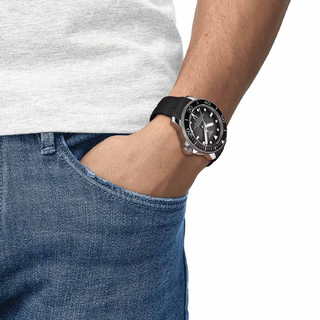 A person in blue jeans and a white shirt stands with their left hand in their pocket, flaunting a Tissot Seastar 2000 Professional Powermatic 80 Automatic 46mm Watch. The elegant black and silver design enhances their style while offering water resistance up to 600 meters.