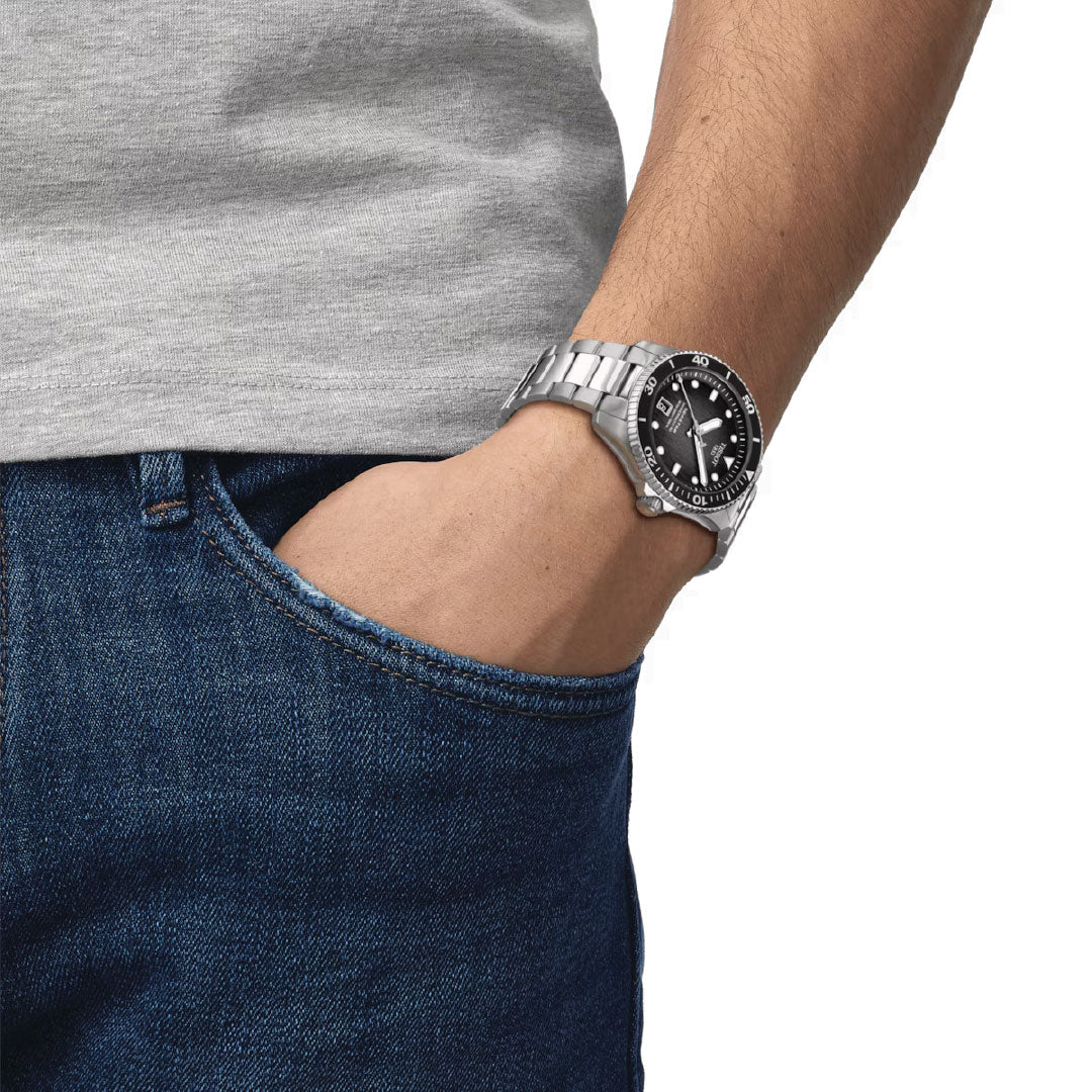 A person wearing a Tissot Seastar 1000 Powermatic 80 Automatic 40mm watch, featuring Super-LumiNova on its black dial, stands with one hand in the pocket of blue jeans. The individual, dressed in a gray T-shirt, exudes casual elegance.