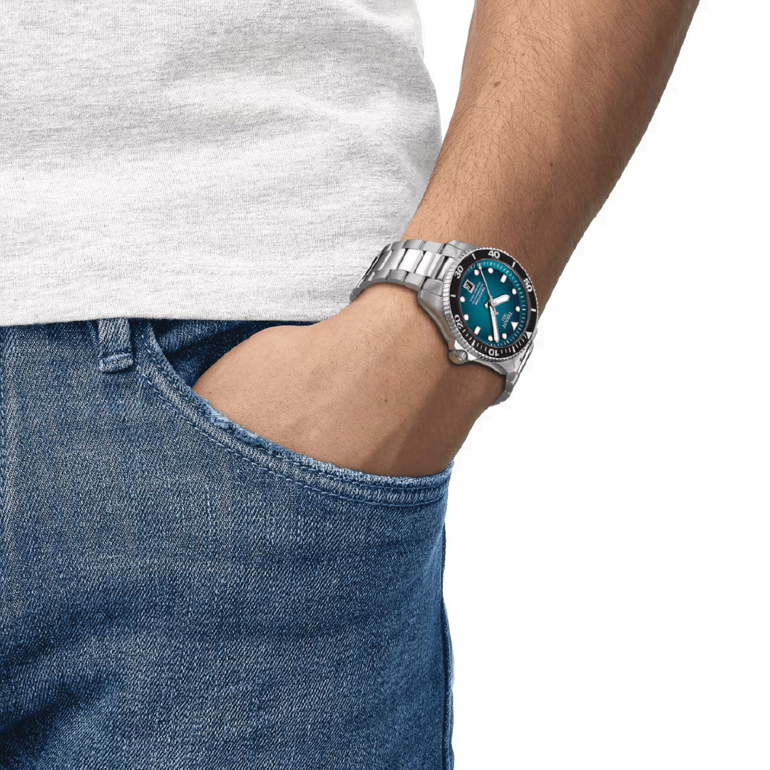 A person casually dressed in blue jeans and a gray shirt has a hand in their pocket. They sport the Tissot Seastar 1000 Powermatic 80 Automatic 40mm Watch, featuring a striking blue face, black bezel, and Super-LumiNova for optimal readability.