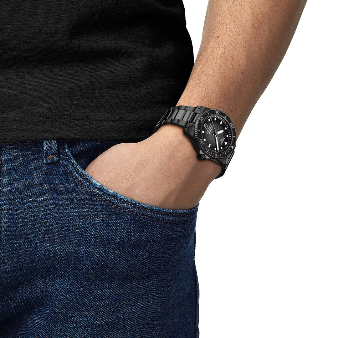 A person in a black T-shirt and blue jeans, left hand in pocket, sports a Tissot Seastar 1000 Powermatic 80 Automatic 40mm watch with a scratch-resistant sapphire crystal and a round face with white details on their wrist.