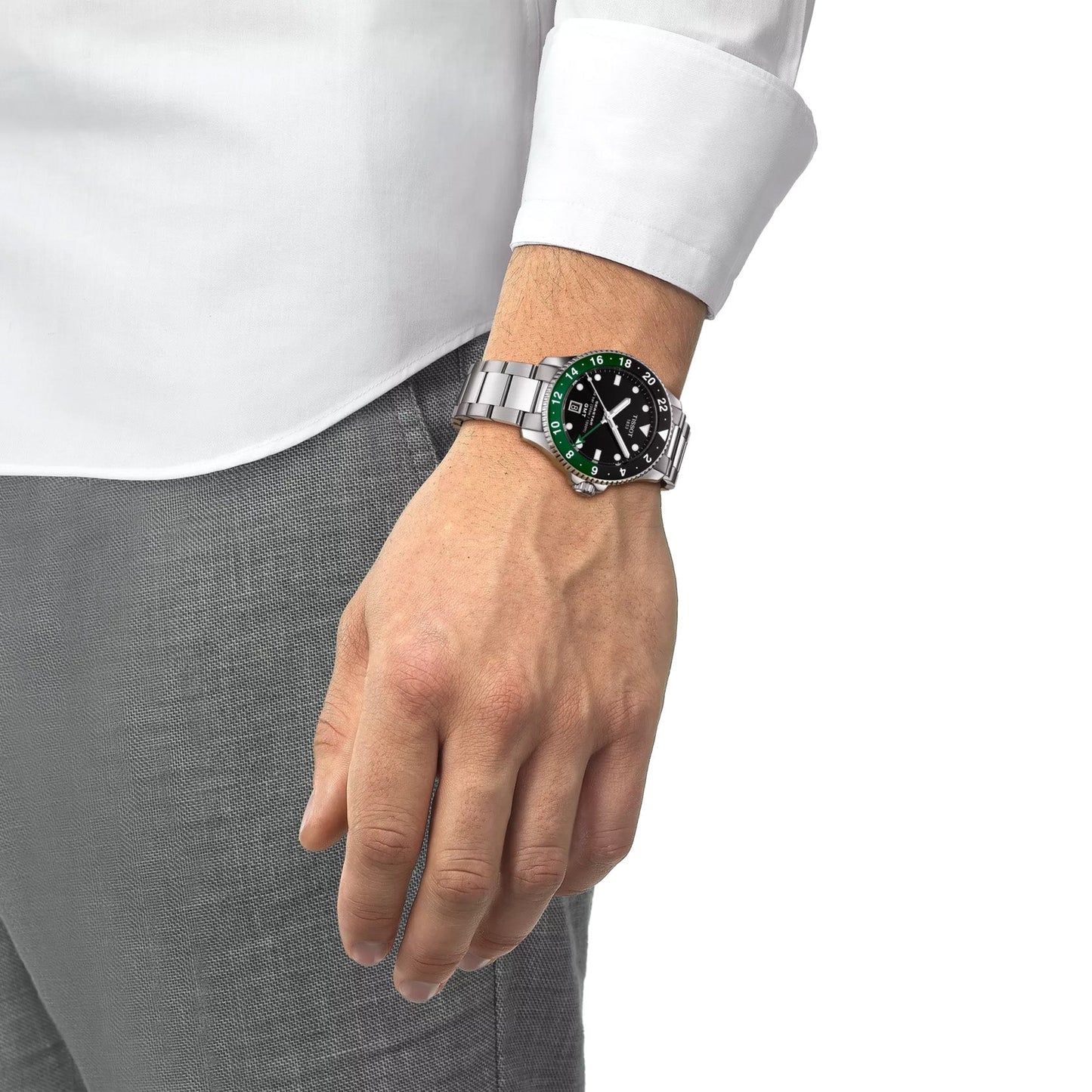 A person wearing a Tissot Seastar 1000 Quartz GMT 40mm Watch, with its green and black face and Swiss Made quartz movement, stands with their hand relaxed by their side. They are dressed in a white shirt and gray pants, perfectly complementing the watch's sophisticated design.