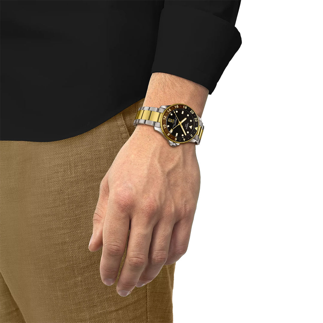 A person in a black shirt and brown pants displays a Tissot Seastar 1000 Quartz GMT 40mm Watch with a black dial on their left wrist, hand relaxed by their side against a white background.