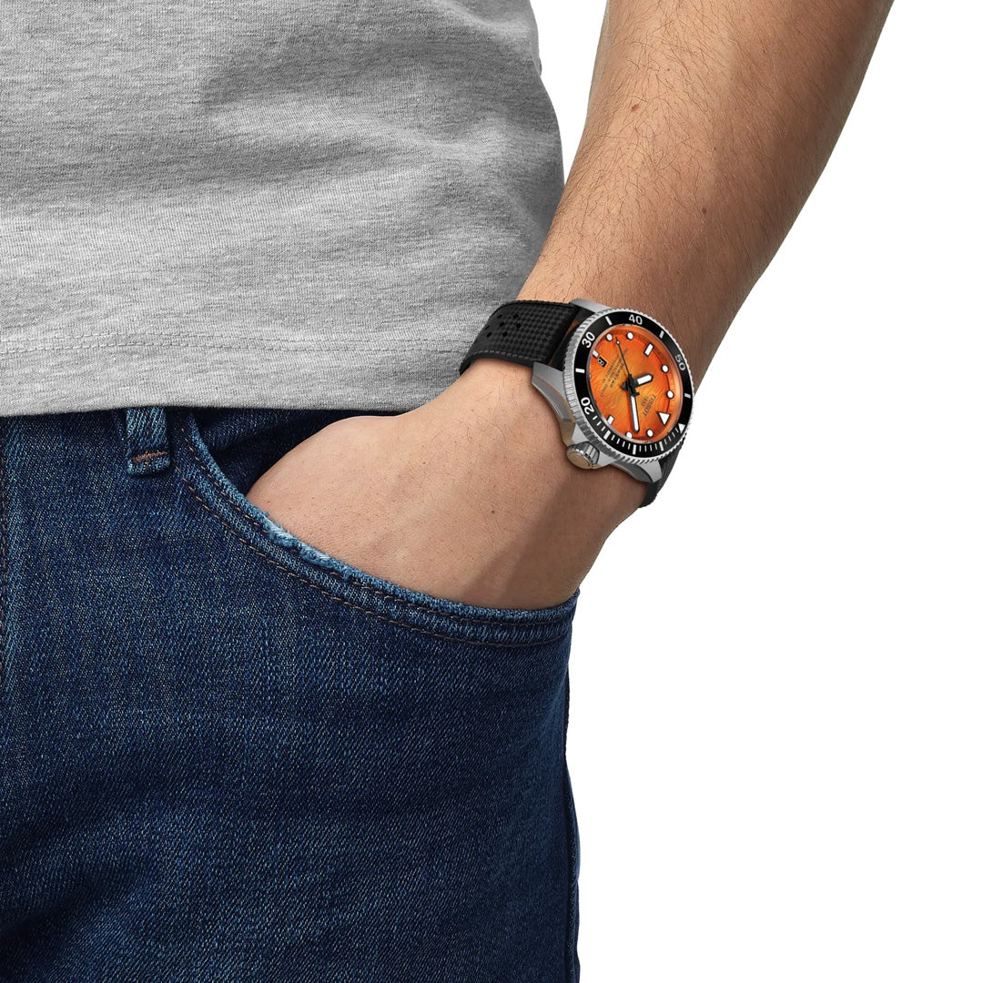 A person wearing blue jeans and a gray shirt has their hand in their pocket, showcasing a Tissot Seastar 2000 Powermatic 80 Automatic 44mm watch with an orange face and black strap.