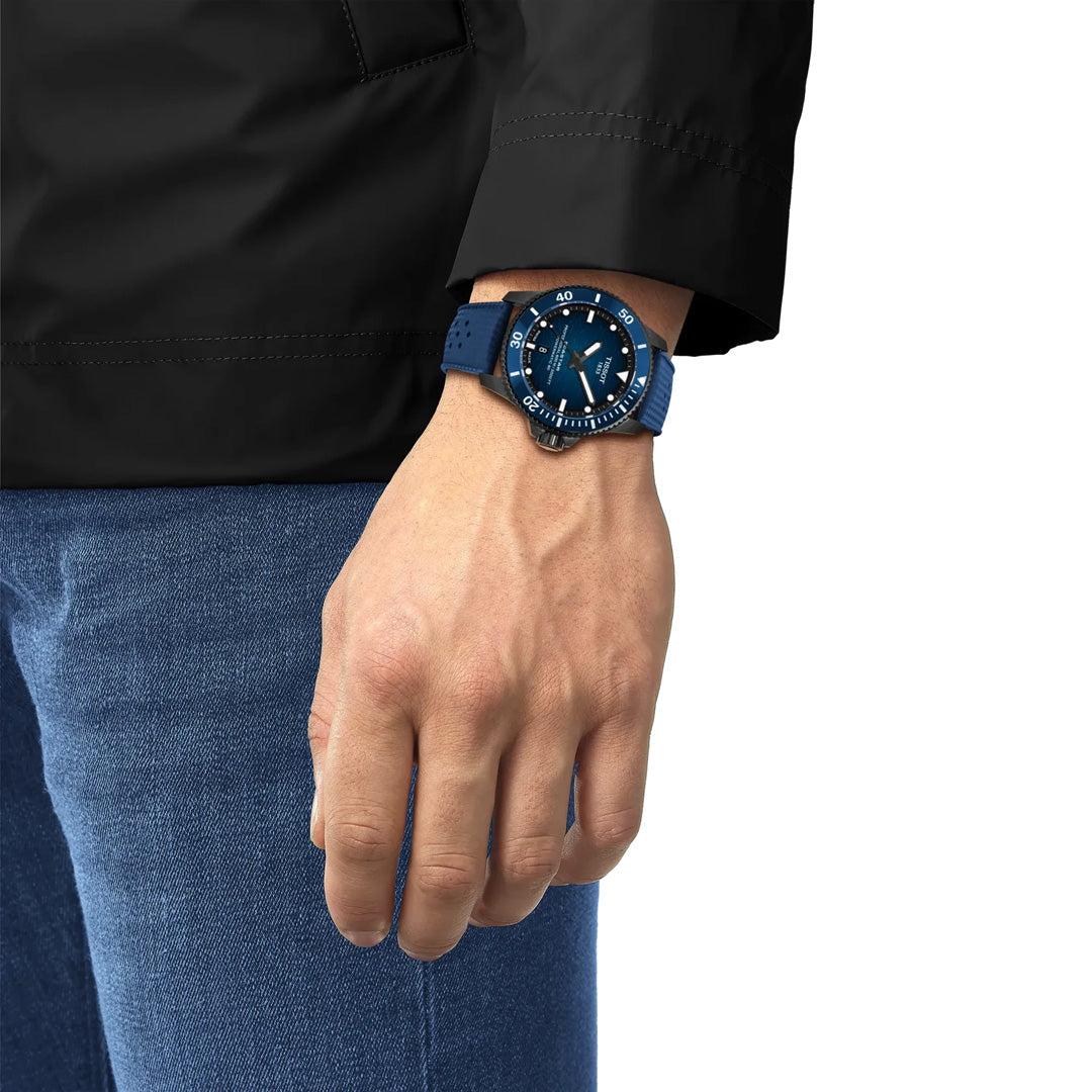 A person dressed in a black jacket and blue jeans displays their left hand and wrist, adorned with the Tissot Seastar 2000 Powermatic 80 Automatic 44mm Watch, its bold round face exemplifying the precision craftsmanship of Tissot’s automatic timepieces.