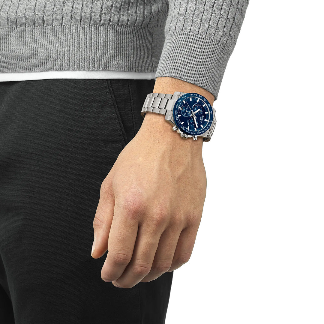 A person wearing a gray sweater and black pants displays the Tissot Supersport Chrono Quartz 42mm watch with a blue face and multiple dials on their left wrist, showcasing this striking Tissot timepiece from Raffi Jewellers.