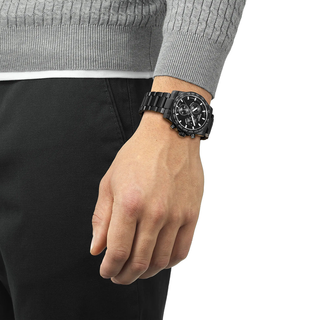 A person dressed in a gray sweater and black pants reveals their left wrist, showcasing a Tissot Supersport Chrono Quartz 42mm Watch with a metal band. With their relaxed hand hanging by their side, they embody the refined elegance associated with Tissot timepieces available at Raffi Jewellers.