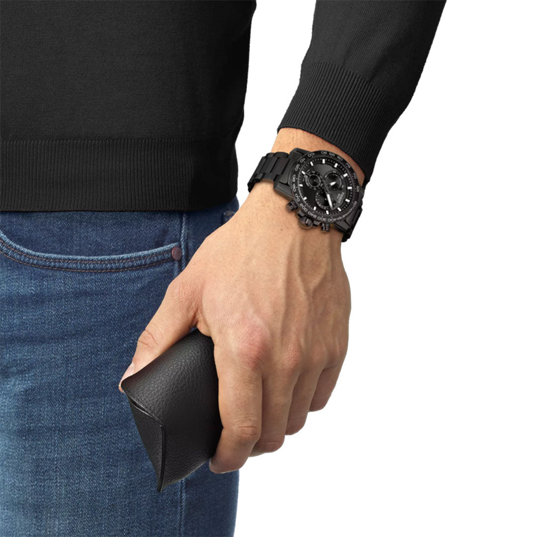 A person in a black long-sleeve shirt and blue jeans holds a black leather case, wearing a Tissot Supersport Chrono Quartz 45.5mm watch by Tissot with a large round dial on the same wrist.