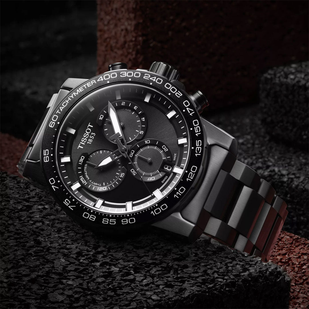 The Tissot Supersport Chrono Quartz 45.5mm Watch by Tissot, featuring a black dial, silver hands, three subdials, and a tachymeter bezel, is displayed on a textured black and red stone surface.