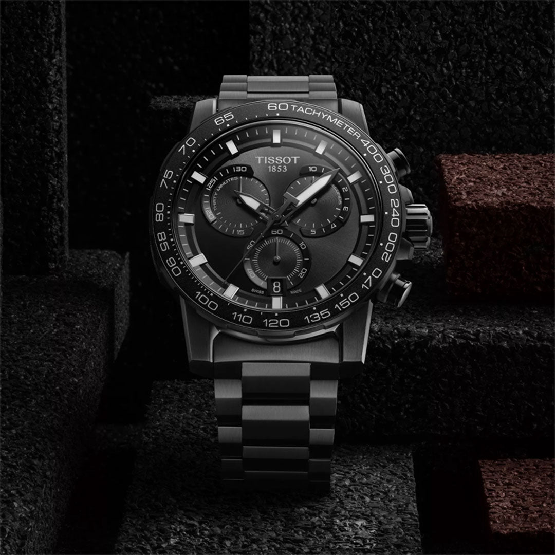 A Tissot Supersport Chrono Quartz 45.5mm watch by Tissot with a black dial and steel case is showcased against a dark, geometric background featuring black and maroon rectangular blocks.