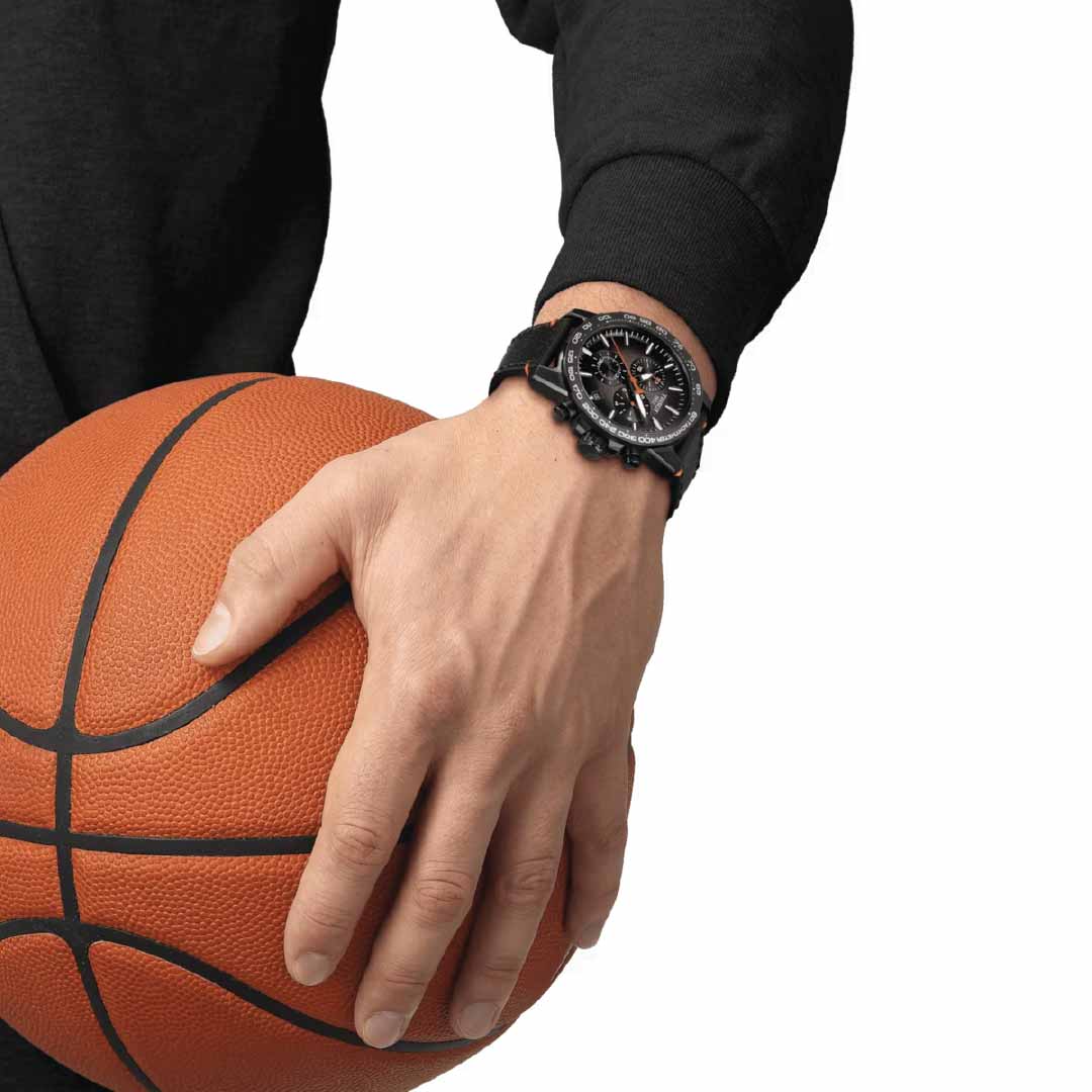 A person wearing a black sweater holds a Tissot Supersport Chrono Basketball Edition Quartz 45.5mm Watch in their left hand. On their left wrist, they sport another Tissot watch, featuring the detailed craftsmanship and precision of Swiss watchmaking against the plain white background.